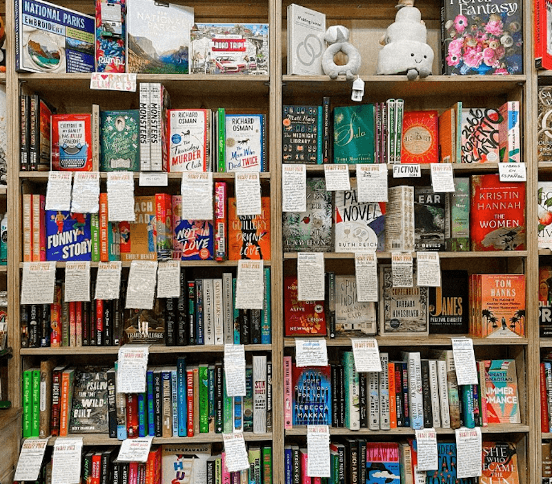 Wooden bookshelf filled with colorful books, each with handwritten staff recommendation notes attached to the covers.