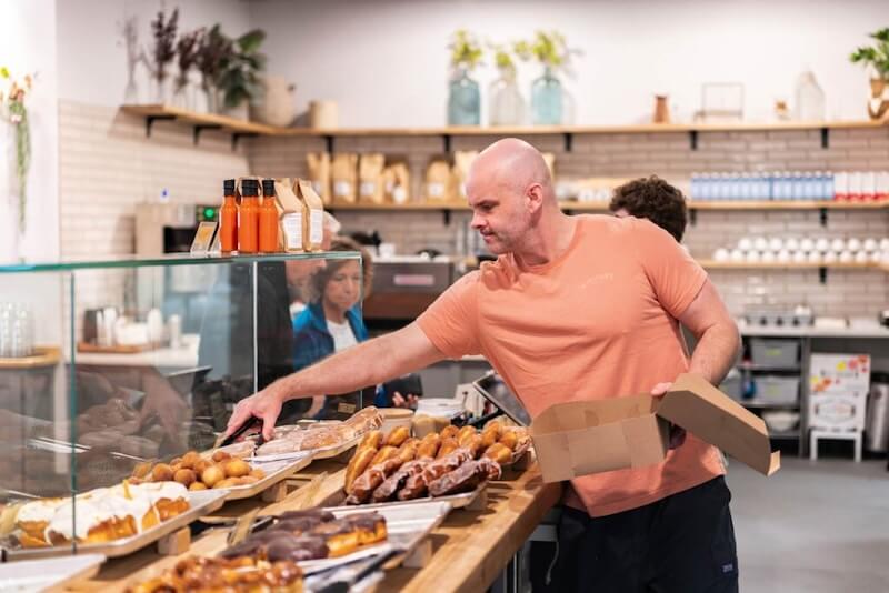 Man in orange shirt selecting pastries from a bakery display, placing them into a box while others wait nearby.