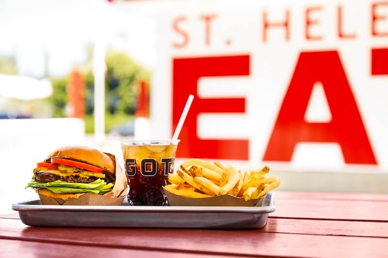Tray with a cheeseburger stacked with lettuce and tomato, a cup of soda with straw, and golden fries on a table.