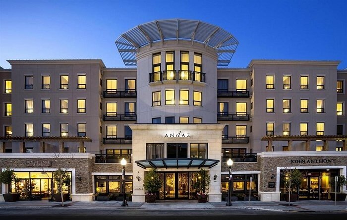 Modern multi-story hotel building lit warmly at dusk, with large windows, balconies, and a central glass entrance.
